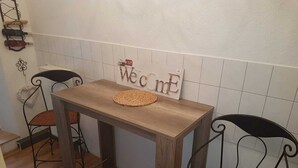 Une table en bois surmontée d’un set de table en fibres tressées, deux chaises en osier et un panneau « Bienvenue » accroché au mur.