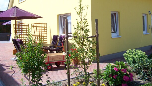 Un edificio amarillo con un patio que tiene una mesa y sillas bajo un paraguas morado, un carrito pequeño y varias plantas.