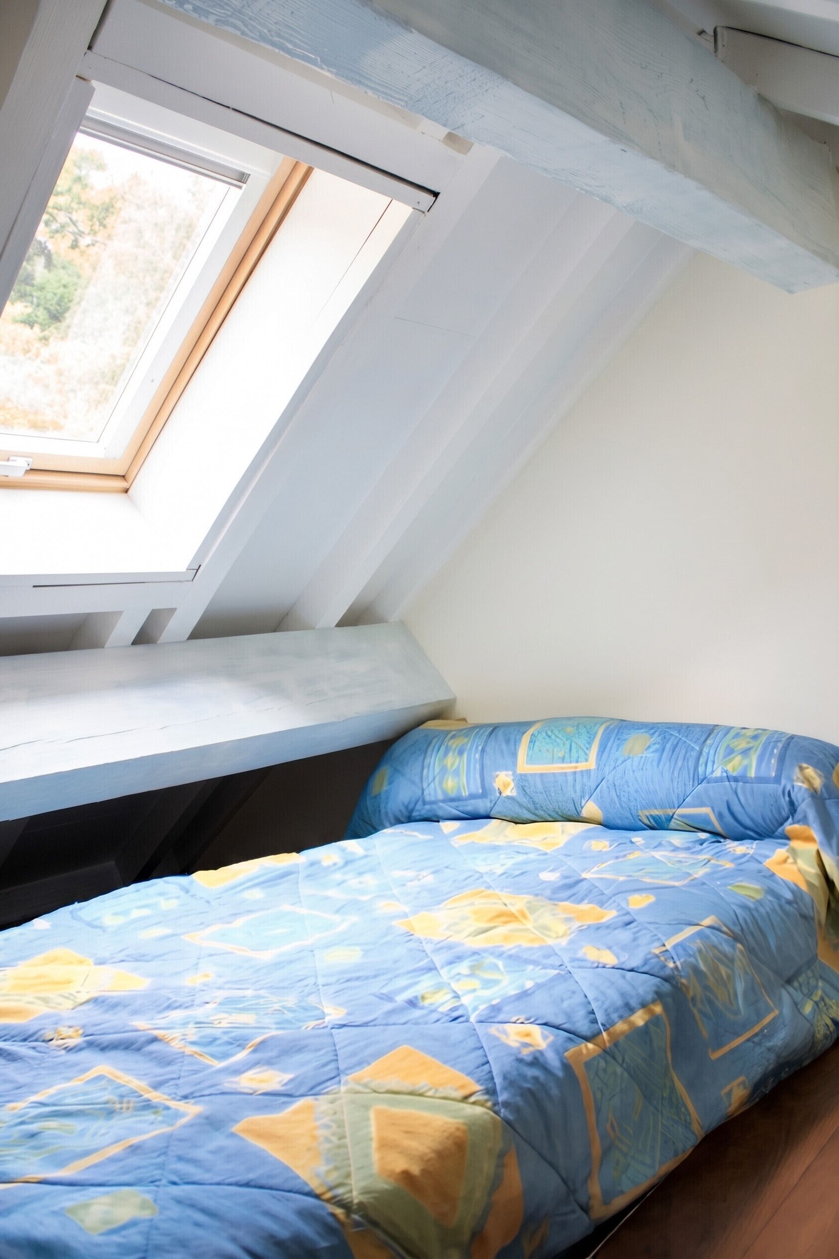 A room with a sloped ceiling, a skylight, a bed with a patterned blue and yellow bedspread, and a wooden floor.