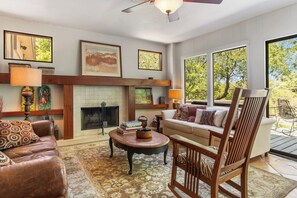 A living room with a fireplace, a sofa, a coffee table, and a chair. There are framed pictures on the wall and a window with a view of trees.