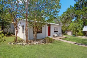 A small white building with a red door, a bench, and a flagpole with an American flag. - Casa del Rio | Pet-Friendly River Stay with Views (Bandera)