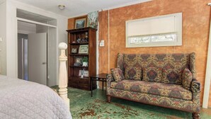 Una habitación con un sofá estampado, una estantería de madera, una puerta que da a otra habitación y una ventana con persianas.