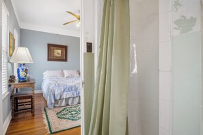Ein Schlafzimmer mit einem Bett, einem Nachttisch, einer Lampe, einem Fenster mit Jalousien, einem Deckenventilator und einem grün gemusterten Teppich.