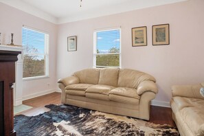A living room with a beige leather sofa, a patterned rug, and two windows with blinds. - Hacienda del Rio | Historic Home on Medina River (Bandera)