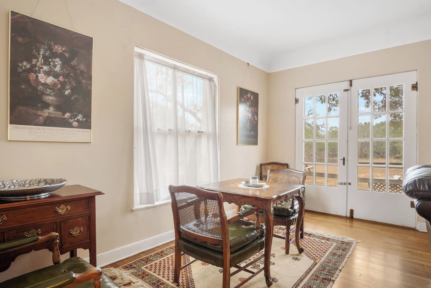 Une salle à manger avec une table et des chaises, une fenêtre ornée de rideaux légers, une porte avec des panneaux de verre et un tapis sur le sol.