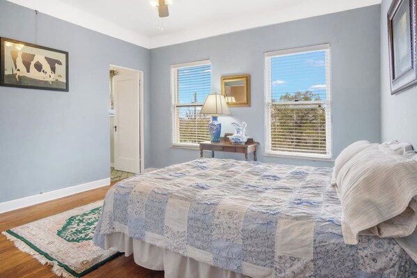 A bedroom with a large bed, a nightstand, a lamp, a mirror, and a window with blinds. - Hacienda del Rio | Historic Home on Medina River (Bandera)