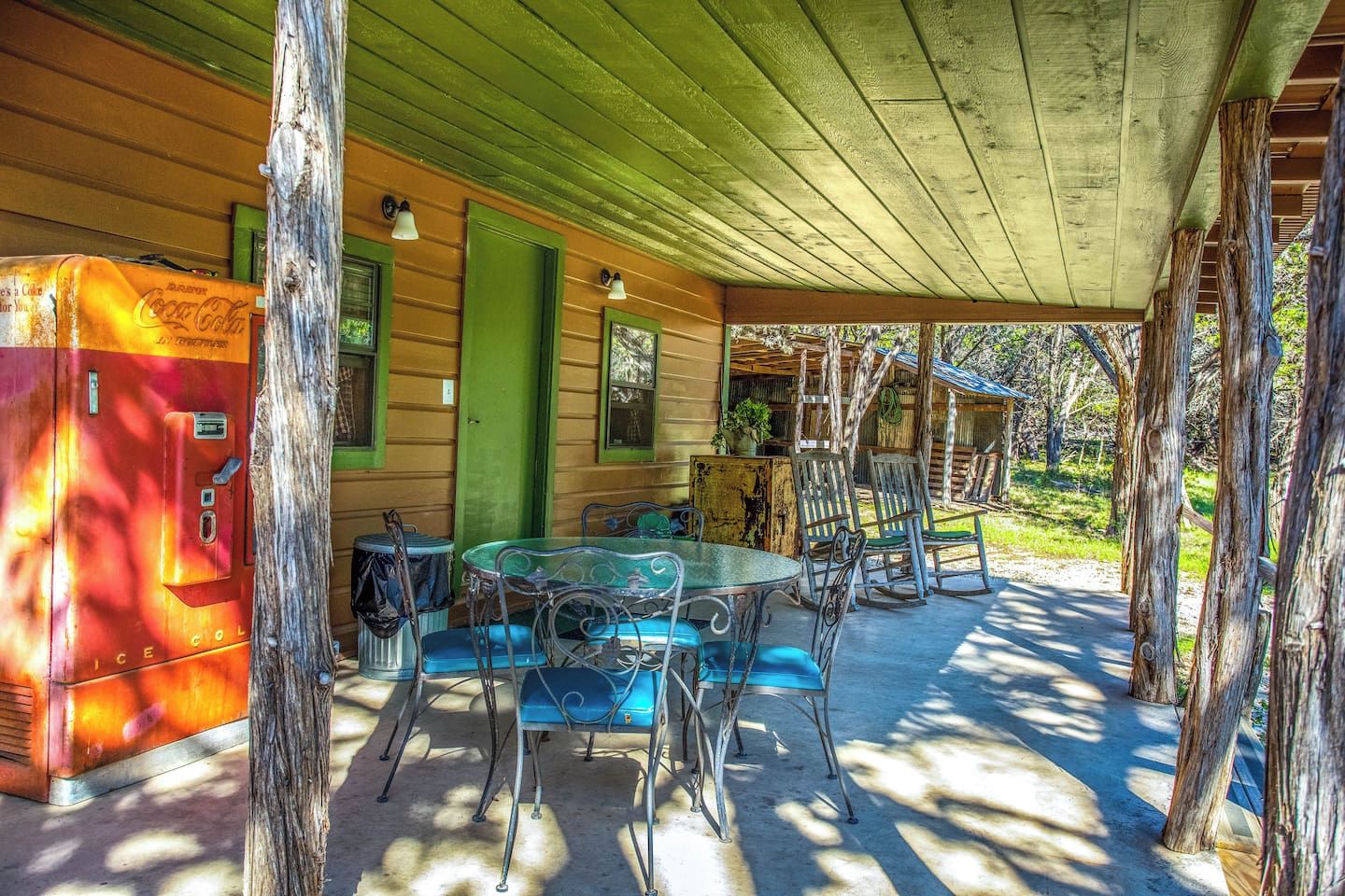 Un espacio al aire libre techado con una mesa, sillas, un refrigerador y una estructura de madera con una puerta verde.