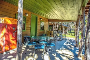 A covered outdoor area with a table and chairs, a refrigerator, and a wooden structure with a green door. - Clear River Cabin | Secluded 2BR w/ River Access (Bandera)