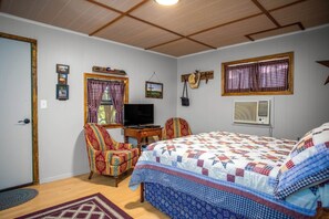 A bedroom with a bed, a chair, a television, and a window with curtains. - Clear River Cabin | Secluded 2BR w/ River Access (Bandera)