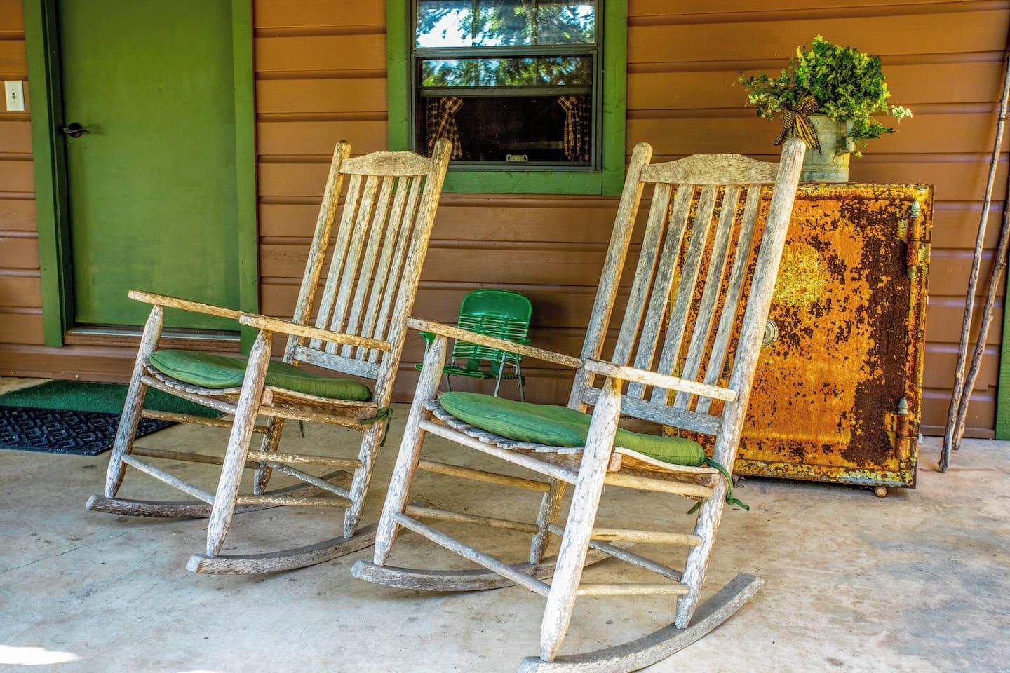 Two rocking chairs with green cushions in front of a wooden house with a green door and a window.