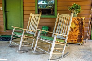 Two rocking chairs with green cushions in front of a wooden house with a green door and a window. - Clear River Cabin | Secluded 2BR w/ River Access (Bandera)