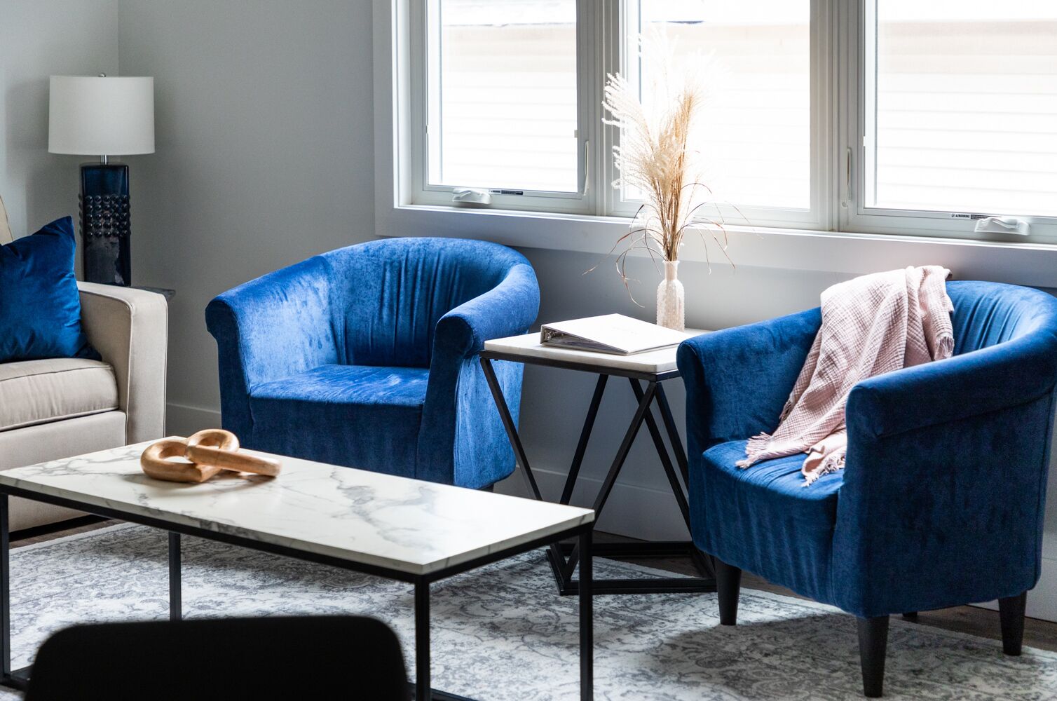 Ein modernes Wohnzimmer mit zwei blauen Sesseln, einem weißen Sofa, einem Marmortisch und einem kleinen Beistelltisch mit einem Buch und einer Vase.