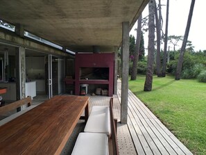 An outdoor dining area with a wooden table and benches, a covered seating area with a red accent wall, and a view of a well-maintained lawn and trees. - Beautiful architecturally designed home, close to everything (Punta del Este)