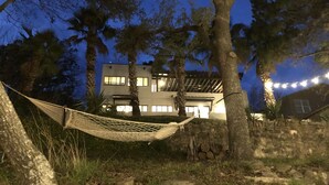 A hammock between trees, a stone wall, and a building with multiple windows and lights at dusk. - Spacious lakefront home with dock - perfect for a group getaway (Lago Vista)