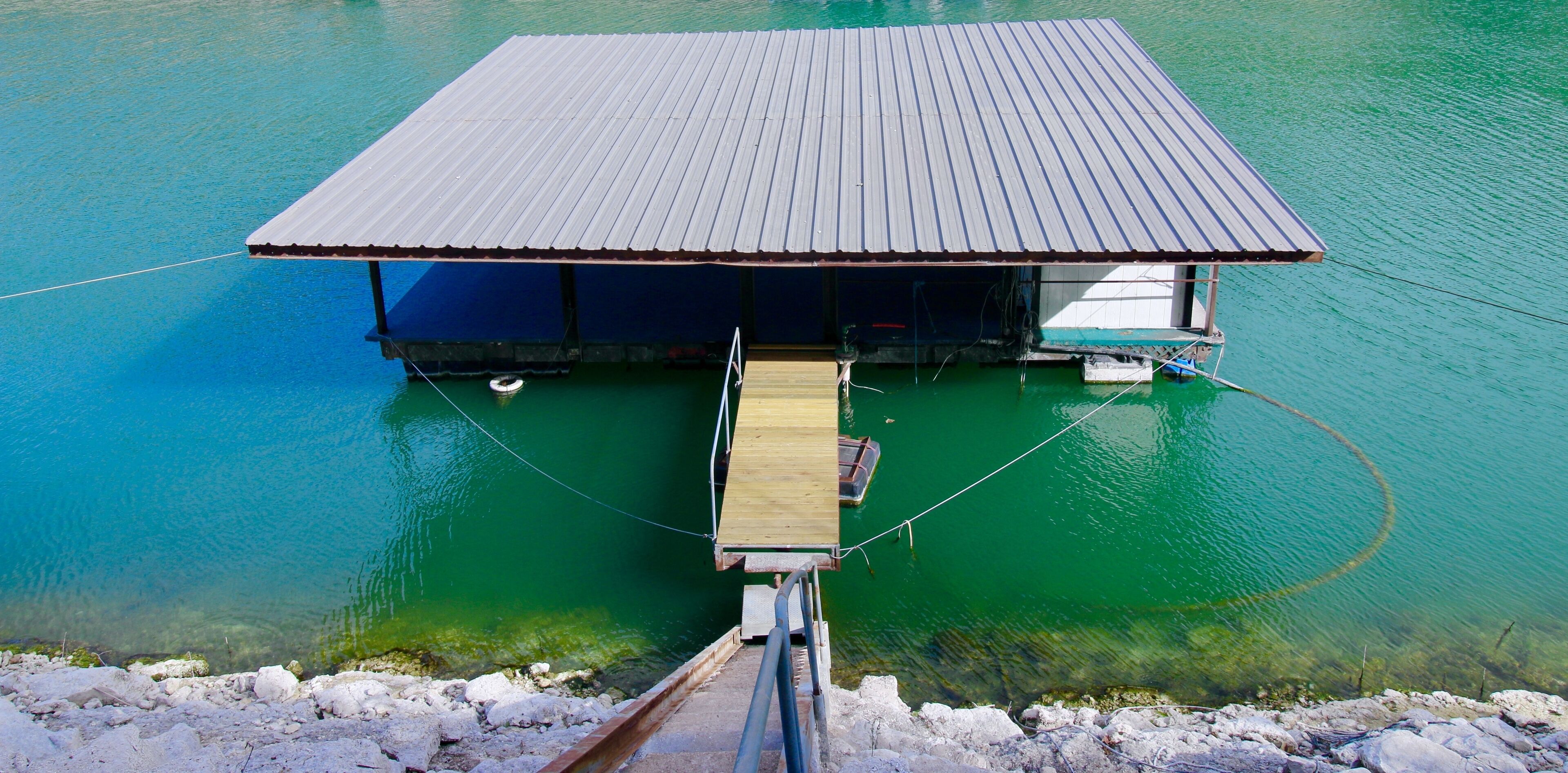 Une structure flottante dotée d’un toit en métal et d’une terrasse en bois qui s’étend au-dessus d’une eau d’un vert cristallin.