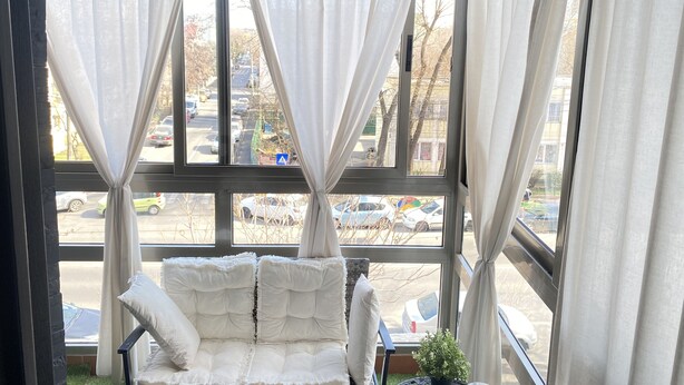 A cozy seating area with a cushioned bench and a small potted plant, overlooking a street through large windows with sheer curtains. - Family Apt ❤️ underground parking❤️smoking balcony (Sector 2)