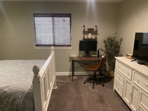 A bedroom with a bed, desk, chair, dresser, TV, window, and a plant.
