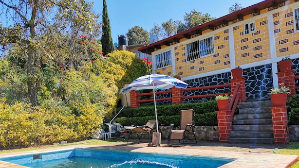 A swimming pool with a tiled deck, surrounded by a garden with various plants and a large building with a brick facade and multiple windows. - Cabaña La Rayuela Valle de Bravo (Valle de Bravo)