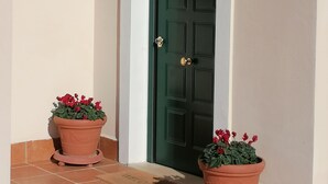 A green door with a brass doorknob, flanked by two terracotta pots with red flowers, and a black wall-mounted lantern above. - Le Vecchie Scuderie (Fara in Sabina)