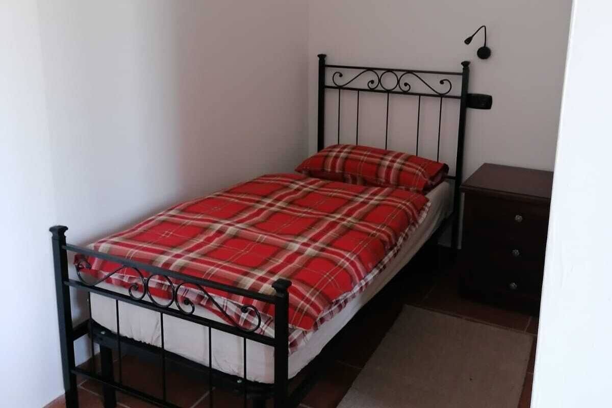 A small, single-bed room with a metal frame, a red plaid bedspread, a wooden nightstand, and a small rug.