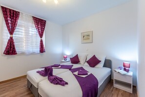 Quarto com duas camas, toalhas roxas, mesa de cabeceira com abajur e janela com cortinas.