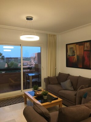 Una sala de estar con un sofá, una mesa de centro con una planta y un cuadro en la pared. Hay un ventanal corredera que da a un área exterior con una mesa y sillas.