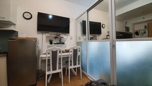 Kompakti keittiö, jossa on jääkaappi, ruokapöytä tuoleineen ja seinälle asennettu televisio.