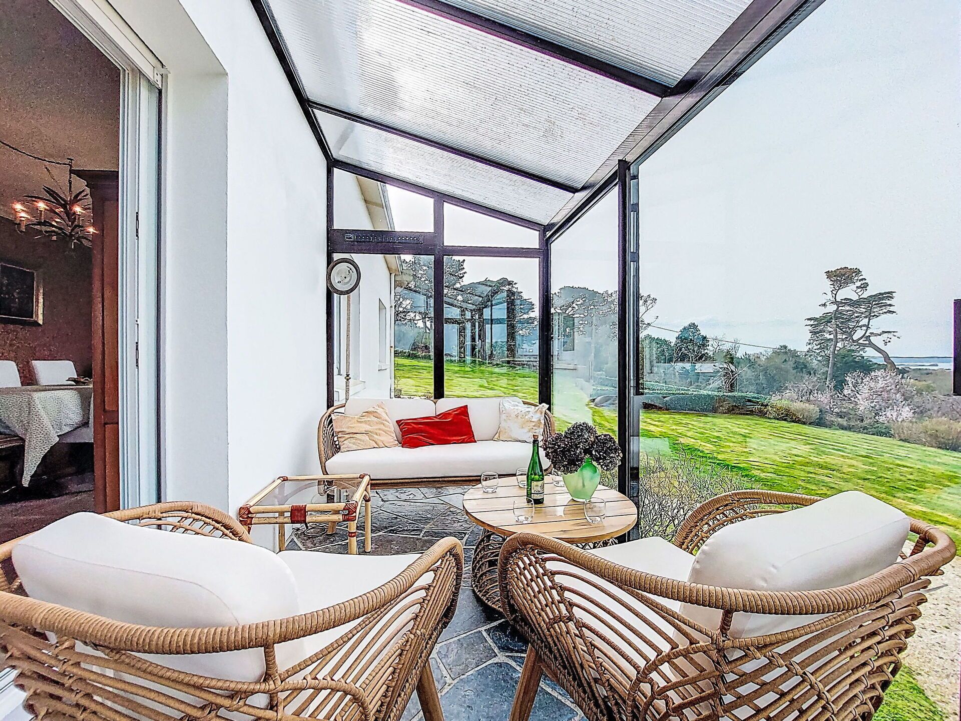 A sunlit patio with wicker furniture, a glass enclosure, and a view of a lush garden.