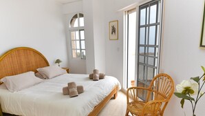 Een slaapkamer met een groot bed, een rieten stoel, een tafeltje met een vaas en een raam met uitzicht.