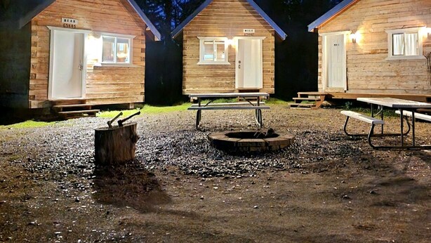 Drie houten hutten met witte deuren, elk met een bankje en een picknicktafel ervoor.