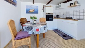 Eine gemütliche Küchenzeile mit Essbereich, mit einem Tisch mit gemustertem Tischläufer, Korbstühlen, einer kleinen Kochinsel mit Kaffeemaschine und Meerblick durch ein Fenster.