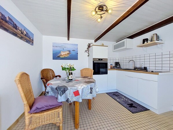 Une cuisine compacte avec un coin repas, comprenant une table recouverte d’une nappe à motifs, des chaises en osier, un petit îlot avec une machine à café et une vue sur la mer par une fenêtre.