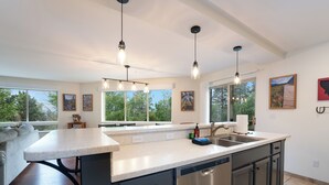A modern kitchen with a large island, pendant lighting, and large windows offering views of the outdoors.