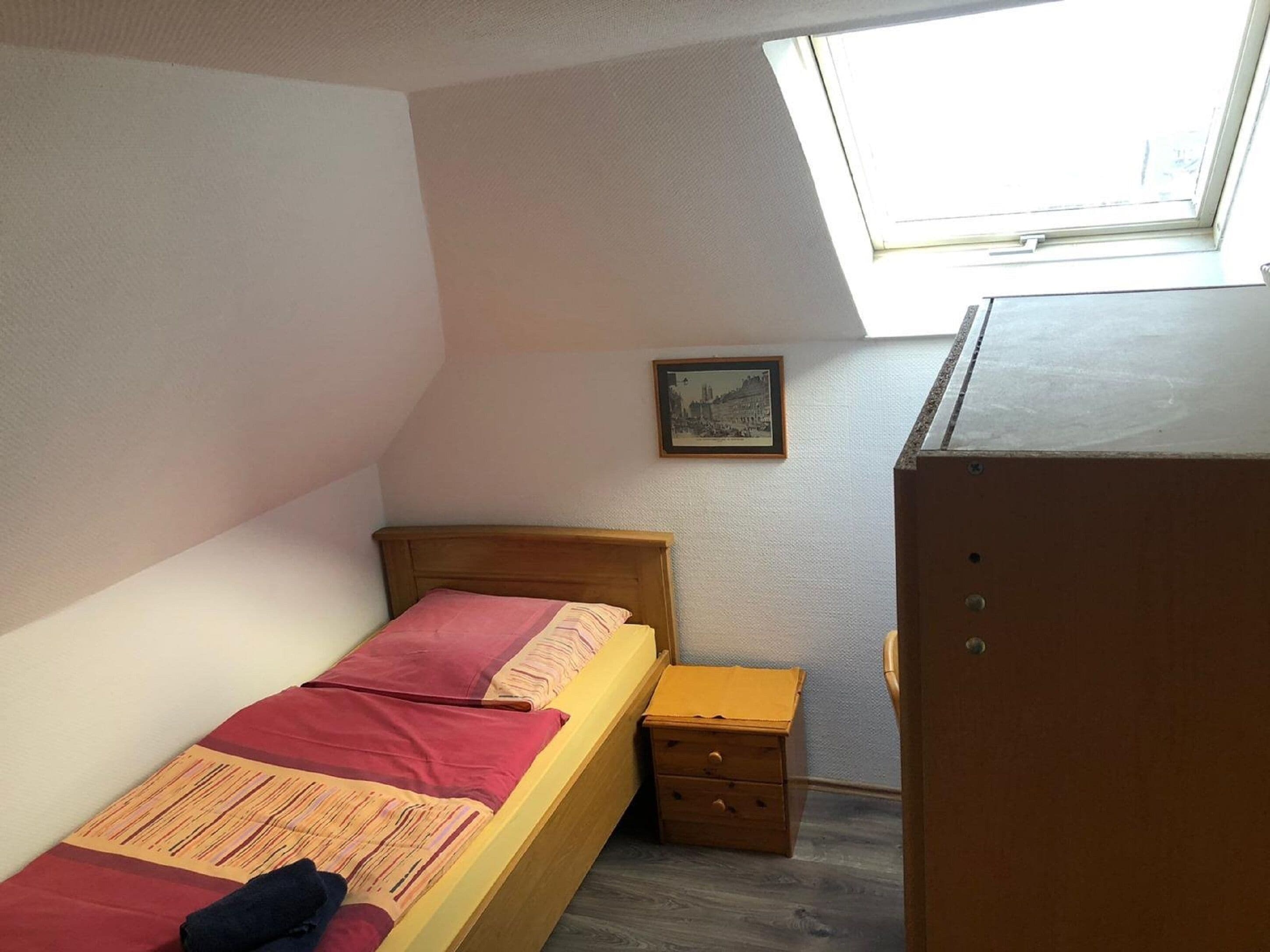 Une petite pièce bien éclairée, avec un plafond en pente, un lit simple doté d’une tête de lit en bois, une petite table de chevet en bois, une commode et un tableau encadré accroché au mur.