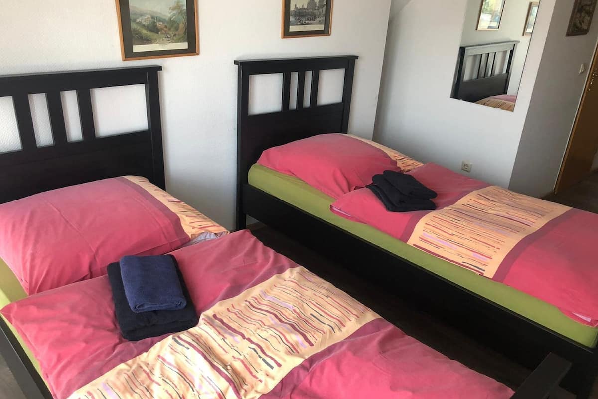 Ein Zimmer mit zwei Betten, jeweils mit Kopfteil und Kissen. An der Wand befinden sich gerahmte Bilder.