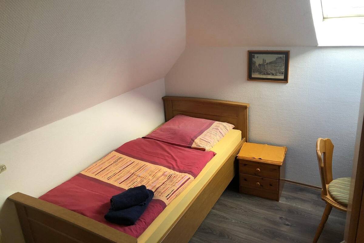 Ein kleines Zimmer mit einem Einzelbett, einem Holzkopfstück, einem kleinen Nachttisch, einem Stuhl und einem gerahmten Bild an der Wand.