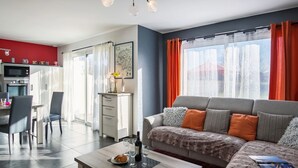 Ein modernes Wohnzimmer mit einem grauen Sofa, orangefarbenen Kissen, einem Couchtisch mit Flasche und Gläsern sowie einem Essbereich mit roter Wand und Küchengeräten.