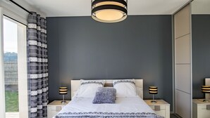 Ein modernes Schlafzimmer mit einem großen Bett, zwei Nachttischen mit Lampen, einer Deckenleuchte und Blick durch ein Fenster mit Vorhängen.