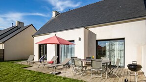 Ein modernes Haus mit einer Terrasse, ausgestattet mit einem Tisch, Stühlen, einem roten Sonnenschirm und einem Grill.