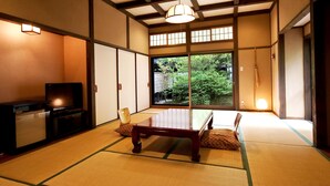 A traditional Japanese room with tatami flooring, a low wooden table, a television, and a view of greenery outside. - Shikinosato HANAMURA (Minamioguni)