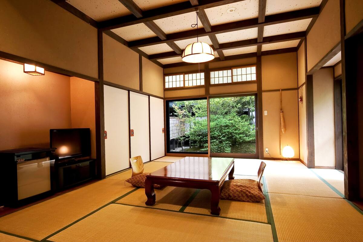 A traditional Japanese room with tatami flooring, a low wooden table, a television, and a view of greenery outside.