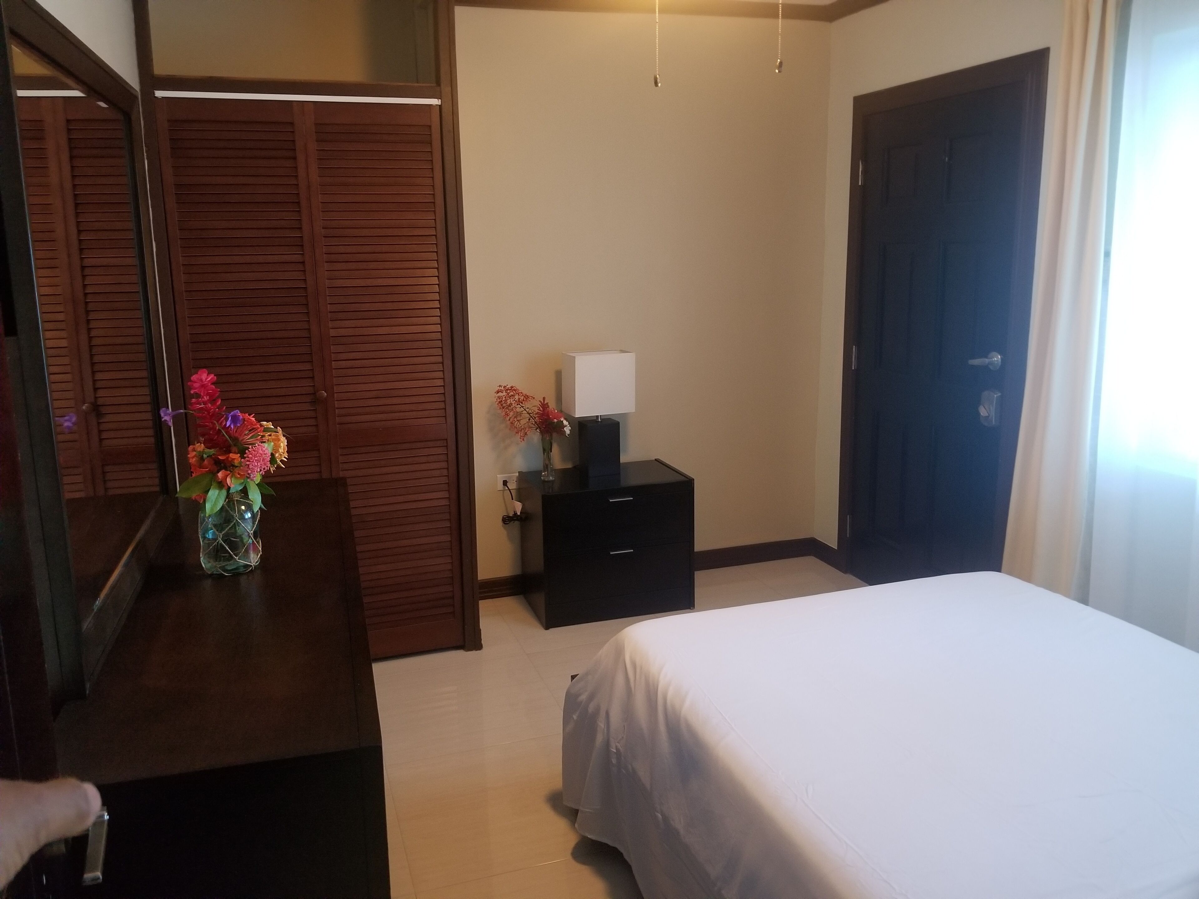 Una habitación de hotel con una cama, un tocador, un espejo, una mesita de noche con lámpara y un armario de madera.