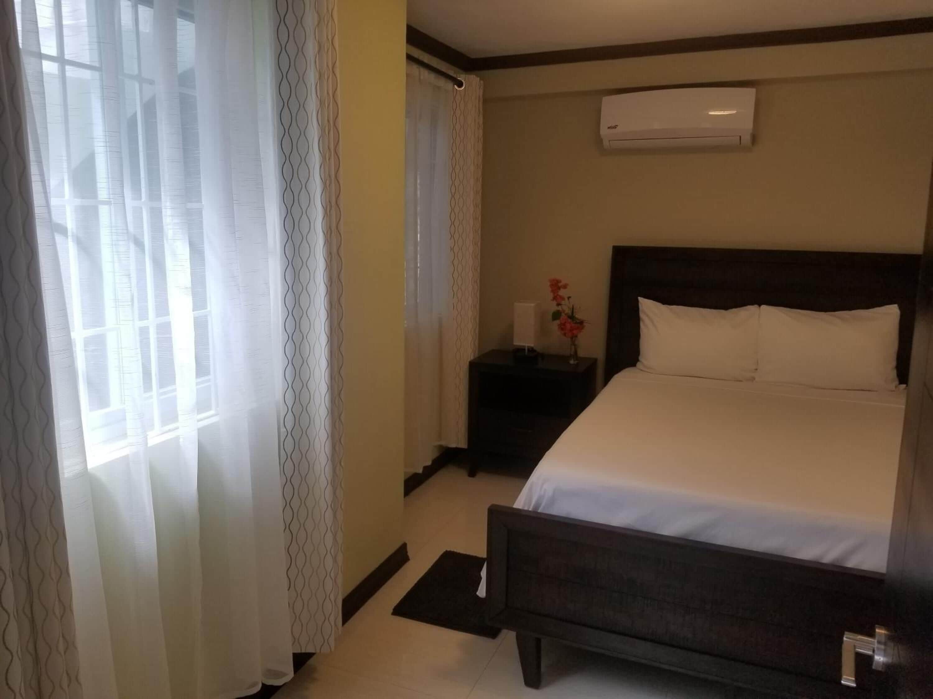 A hotel room with a bed, a nightstand, an air conditioning unit, and a window with curtains.
