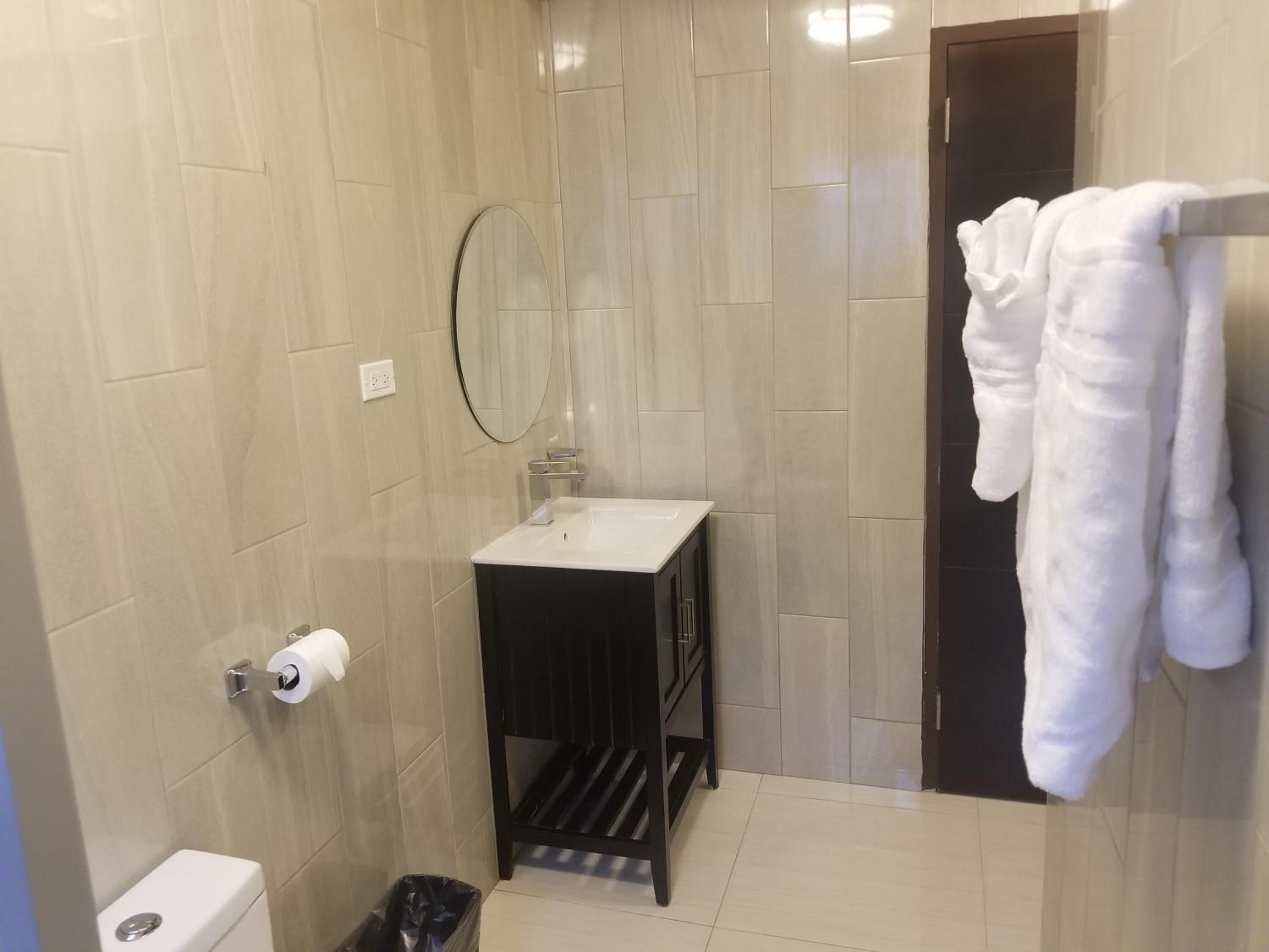 Un baño con baldosas color crema, un lavamanos blanco, un mueble negro, un espejo, un inodoro y un perchero para toallas.