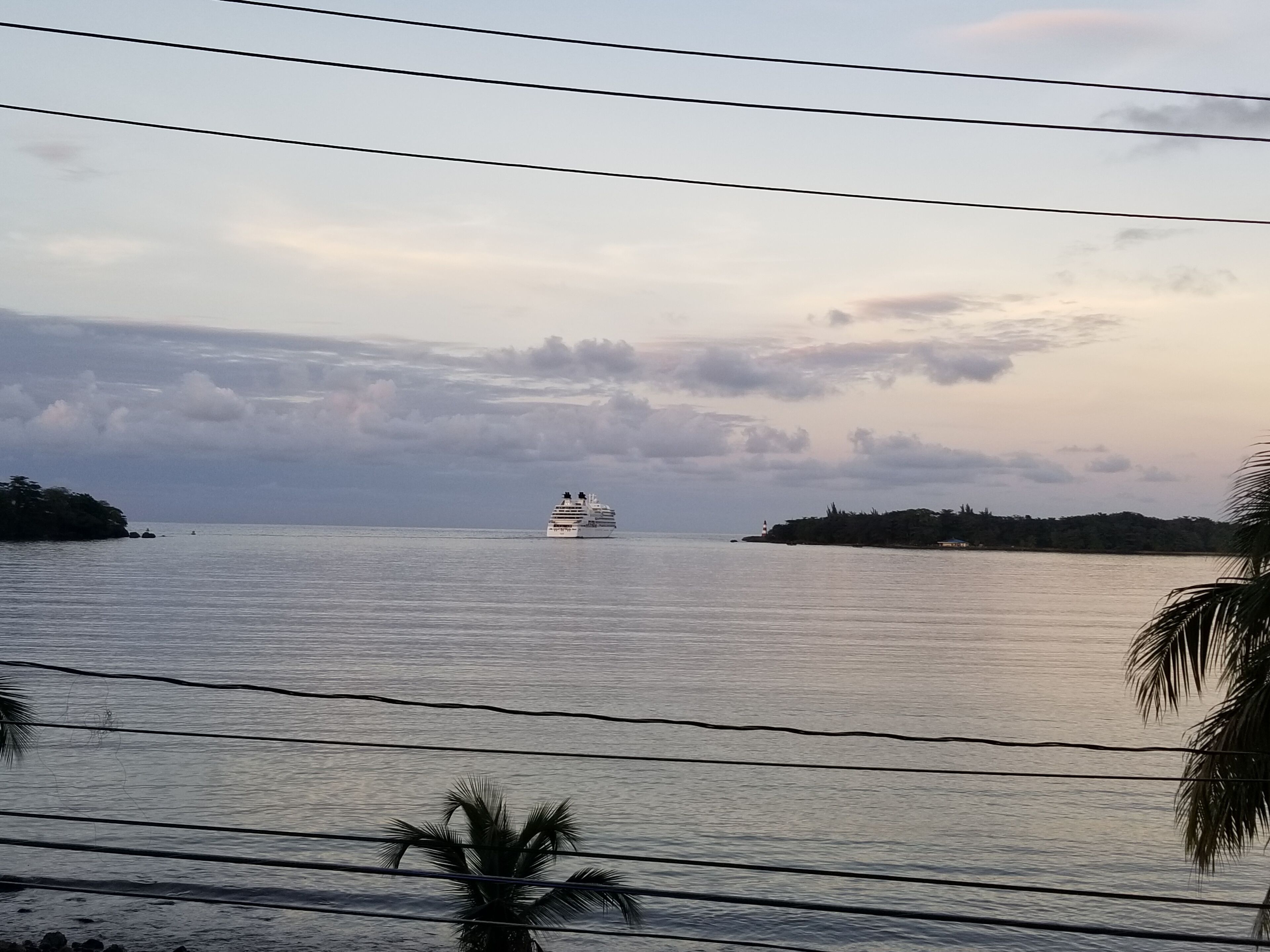A cruise ship on calm waters with a distant lighthouse and a small island with dense foliage.