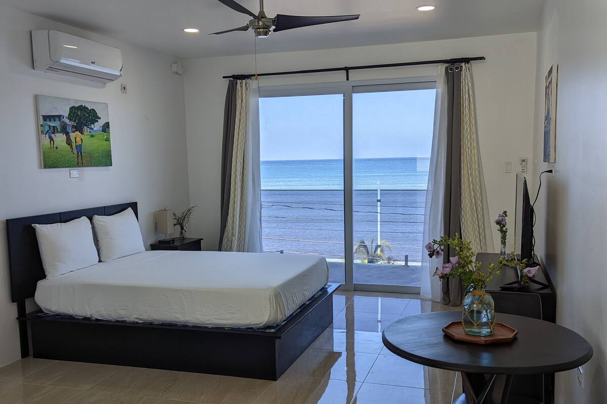 A bedroom with a bed, a desk, a chair, a table, and a view of the sea.