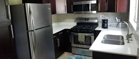 A compact kitchen with dark wooden cabinets, stainless steel appliances including a refrigerator, microwave, stove, and oven, a white countertop, and a small sink.