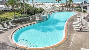 A curved swimming pool with a wooden deck and lounge chairs, surrounded by a white fence, with a beach and palm trees in the background.