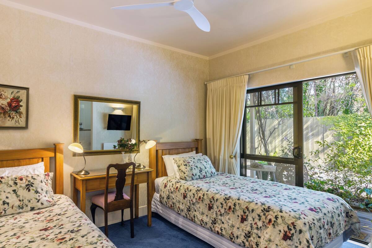 A hotel room with two beds, a desk with a chair, a ceiling fan, and a window with curtains.