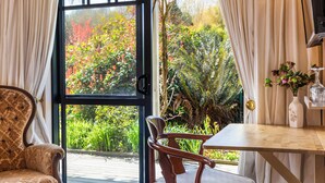 Une pièce avec une table et des chaises en bois, une vue sur un jardin par une porte vitrée, et un vase rempli de fleurs posé sur une étagère.